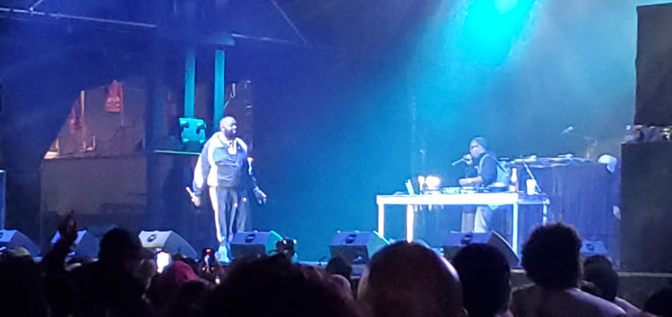 A performer stands on stage with a microphone, while another person is seated at a table with equipment. The stage is lit with blue and purple lights, and there is a crowd in the foreground.
